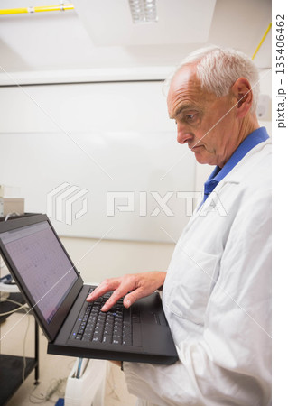 Senior male wearing white lab coat holding laptop typing data in lab by whiteboard, copy space 135406462