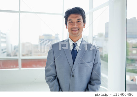Japanese man wearing grey suit and blue tie standing in bright office with floor-to-ceiling windows Japanese man wearing grey suit and blue tie standing in bright office with floor-to-ceiling windows 135406480
