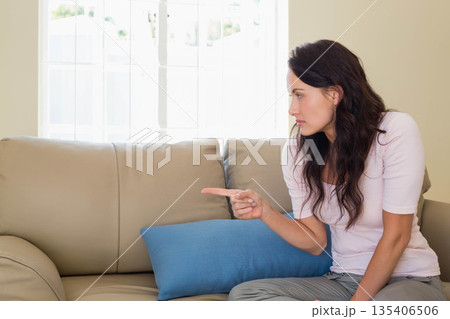 Woman pointing left sitting on sofa in living room with blue cushion near window, copy space Woman pointing left sitting on sofa in living room with blue cushion near window, copy space 135406506