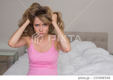 Female pulling hair on bed in bedroom by mirrored nightstand white bedding beige headboard Female pulling hair on bed in bedroom by mirrored nightstand white bedding beige headboard 135406508