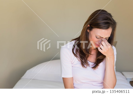Mid adult woman sitting on bed in softly lit bedroom, wiping tears with ring, copy space 135406511