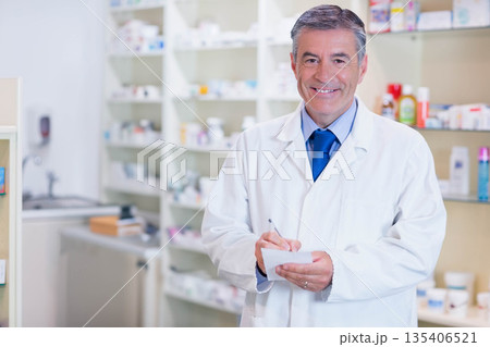 Middle aged male pharmacist writing on notepad with pen at pharmacy desk with shelves, copy space 135406521