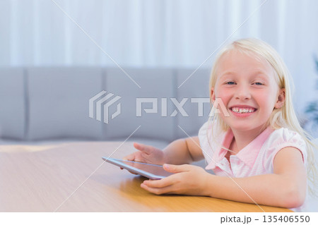 Female child sitting at light wood table inside home holding tablet and smiling, copy space Female child sitting at light wood table inside home holding tablet and smiling, copy space 135406550