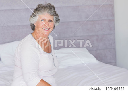 Senior woman sitting on bed smiling in bedroom with white linens and headboard, copy space Senior woman sitting on bed smiling in bedroom with white linens and headboard, copy space 135406551
