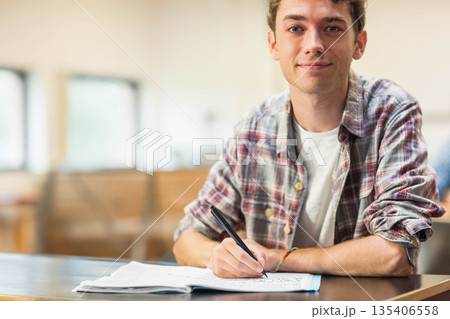 Male student writing notes in open notebook using pen at wooden desk in classroom, copy space Male student writing notes in open notebook using pen at wooden desk in classroom, copy space 135406558