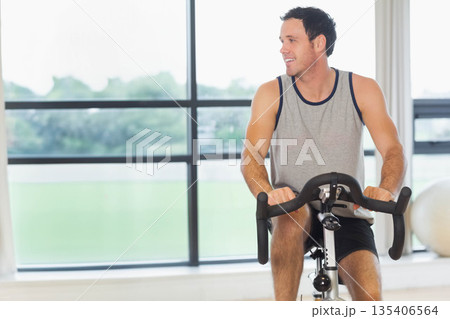 Man in his thirties pedaling bike in gym, exercise ball under floor-to-ceiling windows, copy space Man in his thirties pedaling bike in gym, exercise ball under floor-to-ceiling windows, copy space 135406564