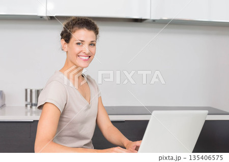 Woman smiling, typing on silver laptop at home kitchen counter with steel canisters, copy space Woman smiling, typing on silver laptop at home kitchen counter with steel canisters, copy space 135406575