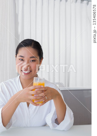 Korean adult female sitting at dining table holding glass of orange juice against curtain backdrop Korean adult female sitting at dining table holding glass of orange juice against curtain backdrop 135406578