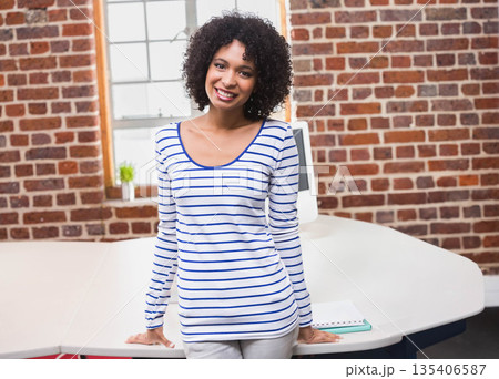 African American woman leaning on white desk in brick office with computer and window smiling African American woman leaning on white desk in brick office with computer and window smiling 135406587