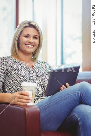 Woman with blonde hair sitting in armchair by tall windows holding paper coffee cup and tablet Woman with blonde hair sitting in armchair by tall windows holding paper coffee cup and tablet 135406592