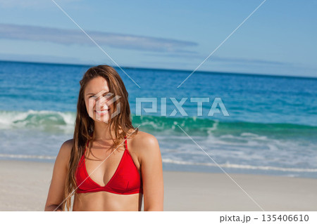 Woman standing on sandy beach at water's edge wearing red bikini top near rolling ocean waves Woman standing on sandy beach at water's edge wearing red bikini top near rolling ocean waves 135406610