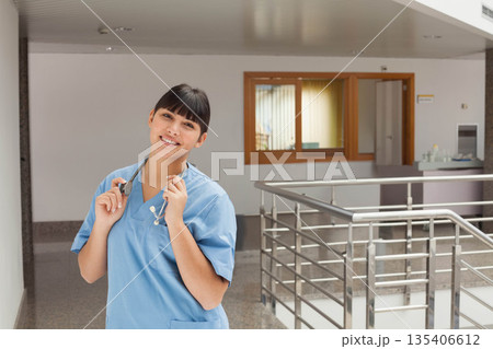 Asian female nurse standing in hospital hall in scrubs holding stethoscope at railing, copy space 135406612