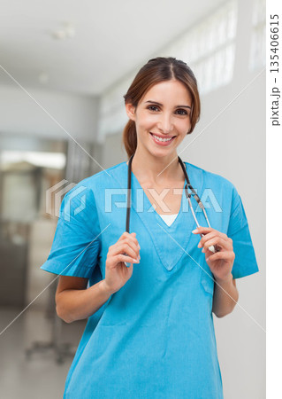 Female nurse in scrubs standing in hospital corridor holding stethoscope around neck smiling Female nurse in scrubs standing in hospital corridor holding stethoscope around neck smiling 135406615