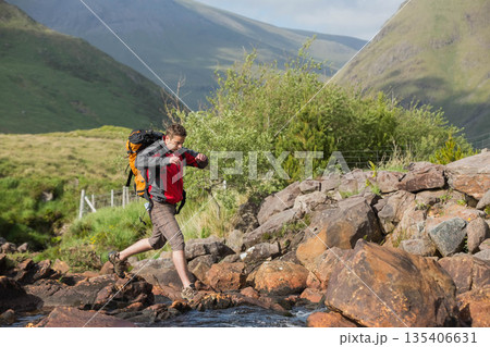 Adult male hiker wearing jacket carrying backpack stepping across stream in valley, copy space Adult male hiker wearing jacket carrying backpack stepping across stream in valley, copy space 135406631