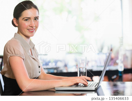Woman sitting at wooden table in bright cafe working on laptop with water glass, copy space 135406633