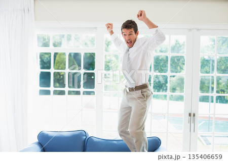 Man in his 30s jumping on blue couch wearing white shirt before grid windows showing pool 135406659