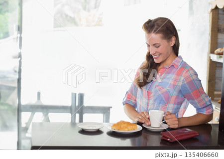Mid-adult woman wearing plaid shirt sitting at café table holding coffee cup, croissant and wallet Mid-adult woman wearing plaid shirt sitting at café table holding coffee cup, croissant and wallet 135406660