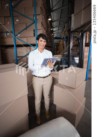 Woman holding tablet and supervising inventory in warehouse in business attire with forklift, boxes Woman holding tablet and supervising inventory in warehouse in business attire with forklift, boxes 135406669