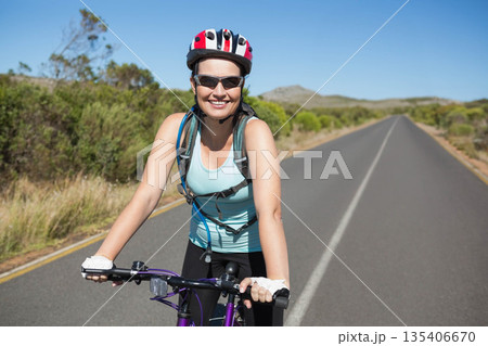 Female cyclist pedaling along rural road wearing helmet sunglasses hydration pack gloves 135406670