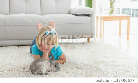Female child lying on ivory rug in living room petting grey rabbits under window, copy space Female child lying on ivory rug in living room petting grey rabbits under window, copy space 135406677