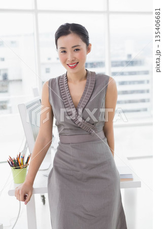 Chinese woman wearing sleeveless dress leaning against office desk reviewing charts on monitor Chinese woman wearing sleeveless dress leaning against office desk reviewing charts on monitor 135406681