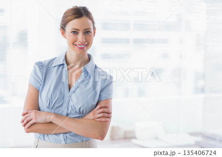 Mid-adult woman crossing arms wearing office attire in office with window blinds and desk 135406724