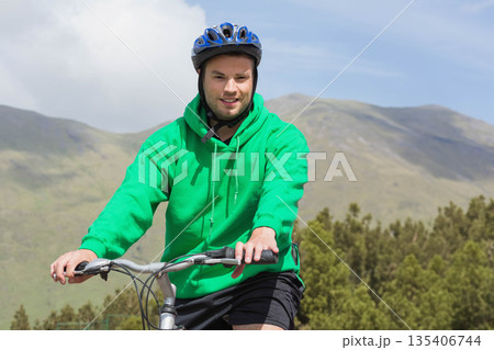 Male cyclist pedaling mountain bike along forest trail wearing green hoodie and blue helmet 135406744