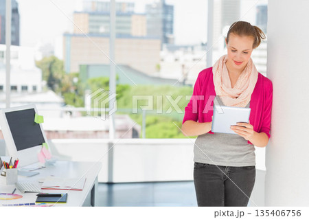 Woman reading tablet device at desk by office windows with computer monitor, copy space 135406756