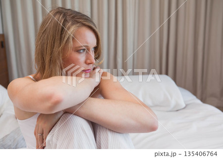 Woman sitting on bed in bedroom hugging knees and gazing right while wearing striped pajama pants Woman sitting on bed in bedroom hugging knees and gazing right while wearing striped pajama pants 135406764