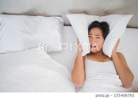 Asian woman clutching pillow over ears in bedroom by beige headboard with white pillows, copy space 135406765