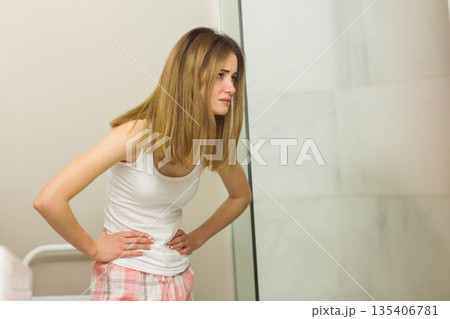 Woman leaning forward in bathroom by shower door wearing sleepwear holding lower abdomen 135406781
