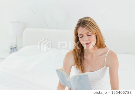 Middle-aged woman sitting on bed reading open book near lamp in white bedroom, copy space Middle-aged woman sitting on bed reading open book near lamp in white bedroom, copy space 135406788