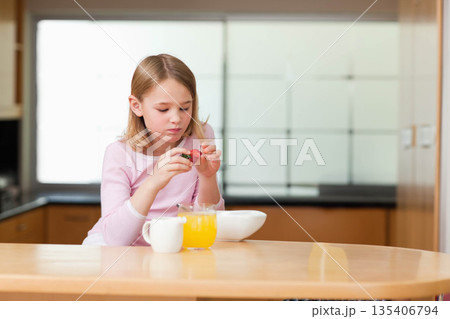 Female child sitting at wood table in home kitchen, holding strawberry above bowl beside pitcher Female child sitting at wood table in home kitchen, holding strawberry above bowl beside pitcher 135406794