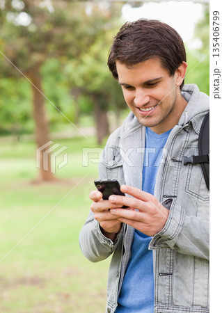 Man standing in park holding smartphone and smiling in denim jacket and backpack, copy space Man standing in park holding smartphone and smiling in denim jacket and backpack, copy space 135406799