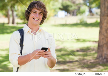 Male student standing in park holding smartphone and wearing backpack in sunlight, copy space 135406803