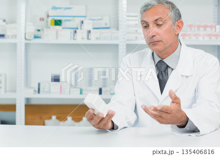 Senior male pharmacist holding medication box while reading label by pharmacy shelves, copy space Senior male pharmacist holding medication box while reading label by pharmacy shelves, copy space 135406816