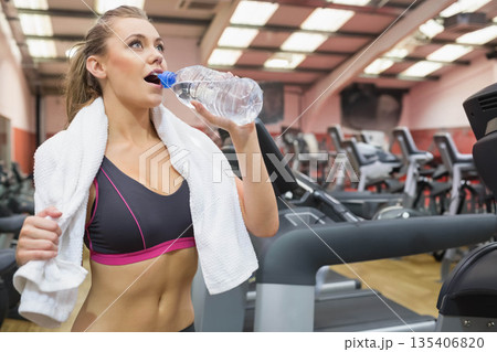 Woman drinking water from clear plastic bottle while pausing treadmill workout in bright gym Woman drinking water from clear plastic bottle while pausing treadmill workout in bright gym 135406820