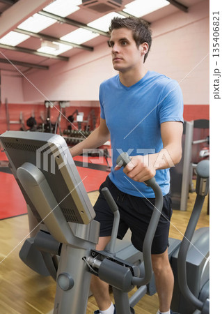 Male wearing blue shirt working out on elliptical trainer in gym amid dumbbells and cardio machines Male wearing blue shirt working out on elliptical trainer in gym amid dumbbells and cardio machines 135406821