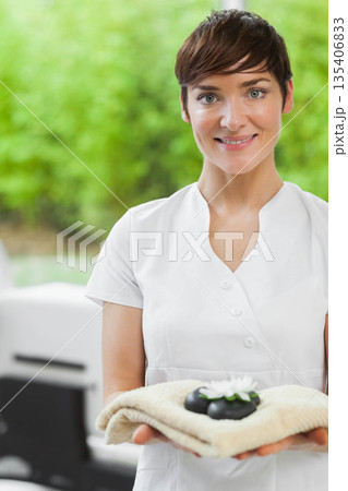 Woman in white uniform holding towel with black stones and white flower in spa, copy space 135406833