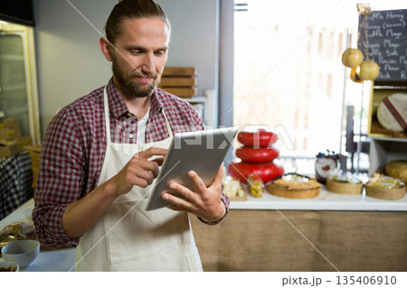 Man using tablet computer behind counter in cheese shop with red-waxed cheese wheels and baskets 135406910