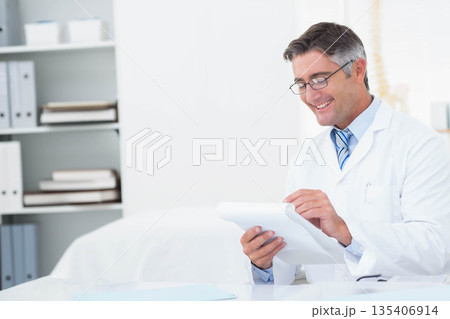 Mature doctor in lab coat reviewing clipboard paperwork at office desk with spine model, copy space 135406914