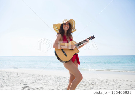 Woman playing acoustic guitar wearing straw hat and red swimsuit on shore beside turquoise ocean 135406923