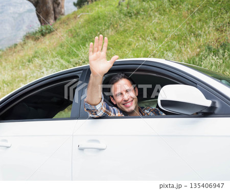 Man wearing plaid shirt waving while leaning out open window of white car at roadside hillside 135406947