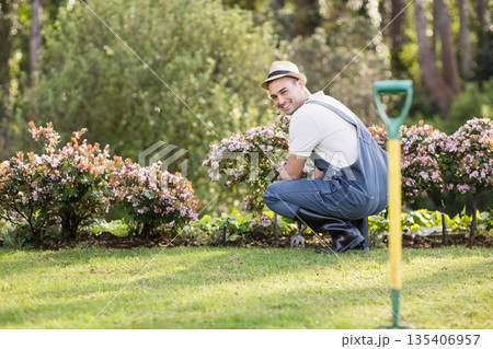Male gardener kneeling in garden tending pink flower bed with shovel and cultivator, copy space Male gardener kneeling in garden tending pink flower bed with shovel and cultivator, copy space 135406957