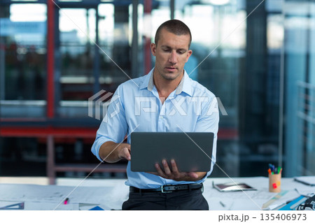 Adult man standing behind desk in modern office typing on laptop among design plans, tablet, pens Adult man standing behind desk in modern office typing on laptop among design plans, tablet, pens 135406973