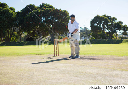 Male cricket player standing on clay wicket gripping wooden bat wearing pads helmet at stumps 135406980