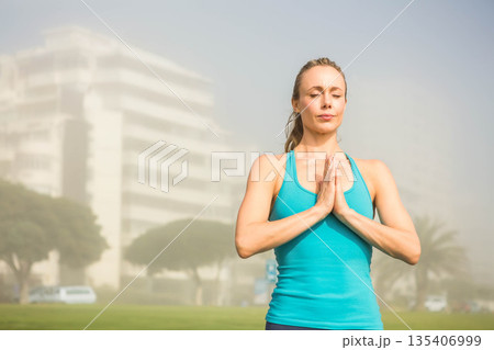 Mid adult female standing in yoga prayer pose on park lawn wearing turquoise sportswear, copy space 135406999