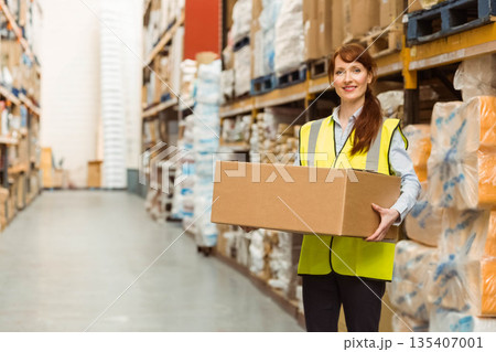 Female worker holding cardboard box standing in warehouse aisle with vest and shelves, copy space 135407001