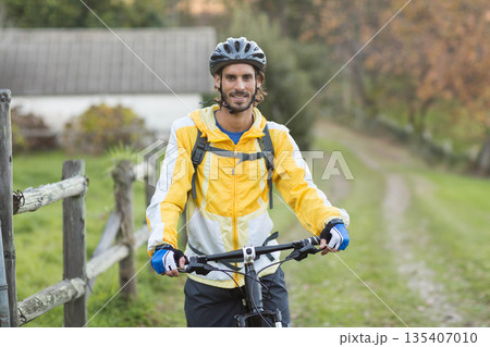 Male cyclist standing beside wooden fence holding mountain bike on rural trail wearing helmet 135407010