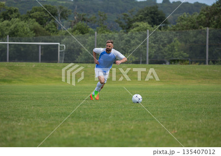 Male athlete sprinting across grass pitch on sports field chasing soccer ball past goal frame 135407012